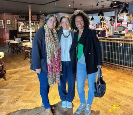 Three women standing together, smiling in a casual indoor setting.