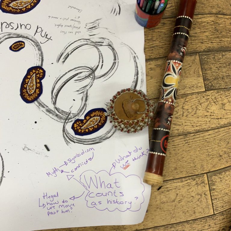 A large piece of  paper with sketches and sentences on it, and next to it are: a maracas, a rain stick and a pot of coloured pens.