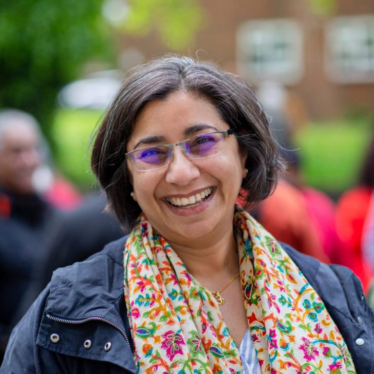 Smiling woman with glasses and a floral scarf, surrounded by blurred figures in the background.